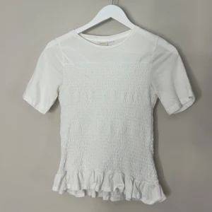 Vit topp med volanger  - Supersöt vit topp med korta ärmar och smockad front och rygg. Toppen har en volangkant längst ner. Perfekt att styla med jeans eller kjol. Materialet känns mjukt och säljer då jag inte använder den, pris kan diskuteras! 