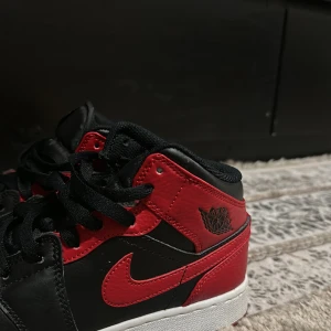 Nike Air Jordan 1 Mid Svart/Röd - Säljer ett par Nike Air Jordan 1 Mid i klassisk svart och röd färg med vita och röda sulor. Skorna har snörning, ikonisk swoosh och Air Jordan-logga på sidan. Tillverkade i skinn med en platt sula och rund tå. Perfekta för dig som vill sticka ut med en tidlös sneaker.