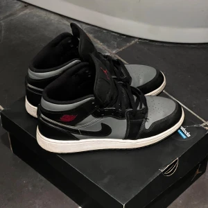 Nike Air Jordan 1 Mid Svart/Grå - Säljer ett par Nike Air Jordan 1 Mid i svart och grått med röda detaljer. Skorna har klassisk snörning, vit sula och svart swoosh. Ovandelen är i skinn och syntet, med ikonisk Jordan-logga på sidan. Mina initsealer står skrivet på lappen i skon men inget som syns.Perfekta för dig som gillar streetwear och sneakers med stil. | för mer frågor och funderingar kontakta mig.✨