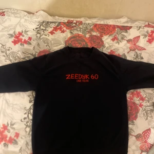 Svart sweatshirt Zeedyk 60 med tryck - Svart sweatshirt från Zeedyk 60 med rött tryck på bröstet och en stor grafisk illustration på ryggen. Tröjan har rund hals och långa ärmar med ribbade muddar. Materialet känns som mjuk bomull och passformen är normal. Perfekt för dig som gillar streetwear med unik design.