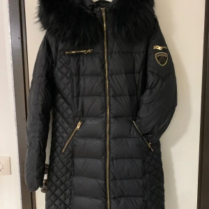 Svart dunjacka Rockandblue med päls - Snygg svart dunjacka från Rockandblue med stor fluffig päls på huvan och guldfärgade dragkedjor. Jackan har quiltade detaljer på sidorna, flera fickor med dragkedja och ett Rockandblue-märke på ärmen. Perfekt för kalla dagar och riktigt stilren look. Jackan och pälsen är äkta.