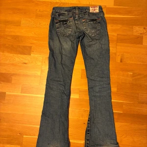 True Religion bootcut jeans blå - Säljer ett par klassiska blå jeans från True Religion med bootcut passform. Jeansen har snygga kontrastsömmar, ikoniska bakfickor med broderad detalj och orangea sömmar. Tillverkade i jeansmaterial och har låg midja. Perfekta för dig som gillar en tidlös och cool look.