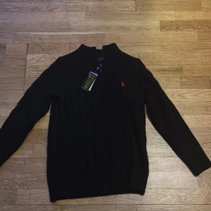 Svart stickad tröja Polo Ralph Lauren - Svart stickad tröja från Polo Ralph Lauren med klassisk kabelstickning och röd broderad logga på bröstet. Tröjan har lång ärm, ribbade muddar och half zip vid halsen. Perfekt för dig som gillar stilren och tidlös design.