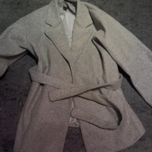 Grå kappa med bälte - Säljer en stilren grå kappa med bälte i midjan och klassisk krage. Jackan har lång ärm och en rak, lite oversized passform. Perfekt för dig som gillar enkel och clean look. Fodrad insida för extra komfort.