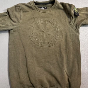 Olivgrön Stone Island sweatshirt - Säljer en olivgrön sweatshirt från Stone Island med stort broderat logotyptryck på bröstet och den klassiska patchen på ärmen. Tröjan har rund halsringning, ribbade muddar och är tillverkad i mjuk bomull. Perfekt för dig som gillar streetwear och snygga detaljer. Knappt använd 