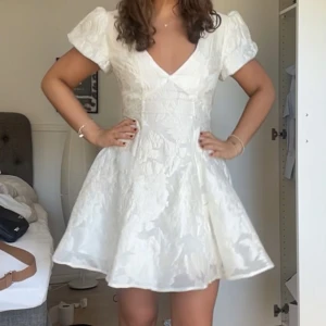 Vit kort klänning med puffärm - Säljer min superfina studentklänning med v-ringning, puffärmar och öppen rygg. Klänningen har ett subtilt blommigt mönster i tyget och en lätt utsvängd kjol. Kunde inte önskat en bättre studentklänning🥰org pris 89 euro