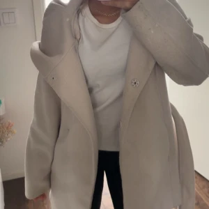 Beige omlott kappa med bälte - Säljer en snygg beige kappa med bred krage och omlottstängning. Jackan har knappar framtill och ett matchande bälte i midjan som ger en clean och stilren siluett. Perfekt för dig som gillar klassisk look med twist. Långärmad och med mjukt material.