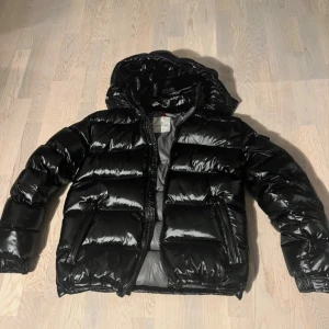 Svart pufferjacka från Moncler - Svart pufferjacka från Moncler med glansig finish och huva. Jackan har två sidofickor med dragkedja och klassisk Moncler-logga på insidan. Perfekt för kalla dagar och har en riktigt snygg streetstil.
