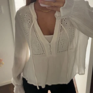 Vit blus med spets och knyt - Säljer en söt vit blus med broderade spetsdetaljer och dekorativa knappar framtill. Blusen har v-ringning, långa ärmar och knytband vid halsen. Materialet är lätt och luftigt, perfekt för varmare dagar. Den har en feminin och romantisk vibe som är superfin till jeans.