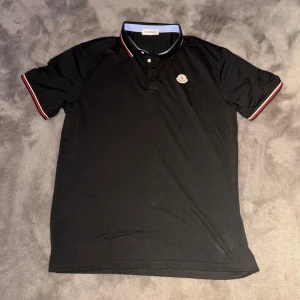 Svart pikétröja från Moncler - Svart pikétröja från Moncler med klassisk krage och knäppning framtill. Tröjan har Moncler-logga på bröstet och röda, vita och blå detaljer på ärmsluten och kragen. Perfekt för en stilren och sportig look.