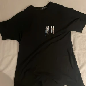 Svart Amiri t-shirt med färgstänk - Svart t-shirt från Amiri med coolt tryck av droppande färg och stor logga både fram och bak.pris kan sänkas vid snabbköp. T-shirten har rund hals och korta ärmar, tillverkad i mjuk bomull. Perfekt för dig som gillar streetwear och vill sticka ut med en unik design.
