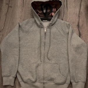 Grå hoodie från Burberry - Grå hoodie från Burberry i storlek M med dragkedja framtill och klassisk huva. Insidan av huvan har Burberrys rutiga mönster. Tröjan har broderad logga på bröstet, snörning vid huvan och två fickor framtill. Perfekt för en chill och stilren look.