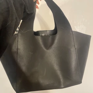 Svart handväska från Carin Wester - Stilren svart handväska från Carin Wester i slätt skinn. Väskan har en enkel och modern design med breda handtag och rymligt huvudfack. Perfekt för dig som gillar minimalistiska accessoarer och vill ha plats för allt du behöver under dagen.