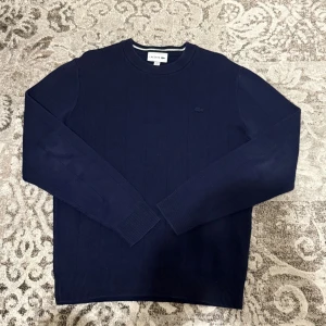 Marinblå stickad tröja från Lacoste - Snygg marinblå stickad tröja från Lacoste med diskret logga på bröstet. Tröjan har rund hals, ribbade muddar och vertikala stickade ränder som ger en clean look. Perfekt för dig som gillar klassisk stil med en modern touch.
