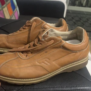 Bruna sneakers från Tommy Hilfiger - Snygga bruna sneakers från Tommy Hilfiger i skinn med vita kontrastsömmar och klassisk logga på hälen. Skorna har snörning och en platt, beige sula som ger en clean och stilren look. Perfekta för dig som gillar en tidlös och casual stil.