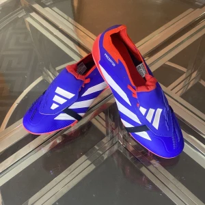 Adidas Predator fotbollsskor  - Säljer ett par Adidas Predator fotbollsskor i blått med vita och svarta detaljer samt röd insida och sula. Skorna har en modern design med tydliga Predator-loggor och mönstrad yttersula för bästa grepp på planen. Perfekta för dig som vill sticka ut på matchen.