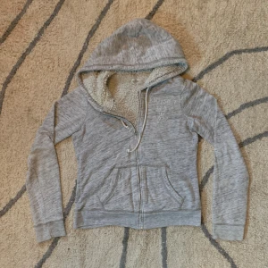 Grå hoodie från Hollister med fuskpäls - Mysig grå hoodie från Hollister med broderad logga och årtal på bröstet. Tröjan har huva med snörning, dragkedja och två fickor framtill. Insidan är fodrad med fluffig fuskpäls som gör den extra varm och skön. Perfekt för kyliga dagar! Den är i bra skick men sitter lite litet i passformen