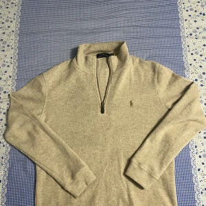 Beige half zip tröja Polo Ralph Lauren - Snygg beige långärmad tröja från Polo Ralph Lauren med half zip och klassisk broderad logga på bröstet. Tröjan har ribbade muddar och är gjord i mjuk bomull, perfekt för chill dagar. Passar dig som gillar stilren och enkel design.