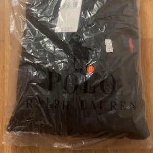 Marinblå stickad tröja Polo Ralph Lauren - Säljer en marinblå stickad tröja från Polo Ralph Lauren med klassiskt kabelstickat mönster och röd broderad logga på bröstet. Rund halsringning och ribbade muddar. Perfekt för dig som gillar stilrena och tidlösa plagg.