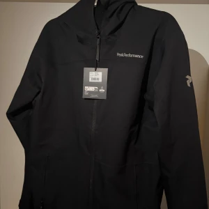 Svart vindjacka Peak Performance S - Peak Performance svart vindjacka med huva och hel dragkedja framtill. Snyggt broderad logga på bröstet och diskret logotyp på ärmen. Jackan har en clean look och är tillverkad i slitstark polyester, perfekt för dig som gillar stilren och funktionell design.