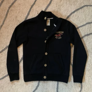 Holliste overshirt/jacka  - Svart kofta från Hollister med broderat logotyp och text 'SO CAL' på bröstet. Tröjan har ribbade muddar, ståkrage och stängs med både dragkedja och stora knappar. Fickan till höger är lite slitet. Materialet är mjukt och känns som bomull, perfekt för kyliga dagar.