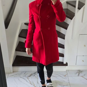 Röd kappa med dubbelknäppning - Snygg röd kappa med figurnära passform och hög krage. Dubbelknäppning framtill med stora knappar och fickor på sidorna. Klassisk och stilren modell som ger en riktigt clean look. Perfekt för dig som vill sticka ut i höst och vinter.