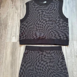 Svart och grå kortkjol samt väst från H&M - Cool kortkjol från H&M i svart och grått med ett hypnotiskt rutigt mönster. Kjolen har en ribbad midja och är gjord i mjukt stickat material som känns skönt mot huden. Perfekt att matcha med denna crop väst för en trendig look. Kjolen är stl S och västen stl M.