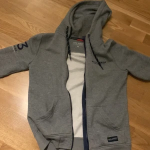 Grå hoodie från Race Marine XS - Grå hoodie från Race Marine i storlek XS med dragkedja och huva. Tröjan har svarta snören med vita detaljer, fickor fram och tryck på ena ärmen. Insidan är mjuk och fleecefodrad, perfekt för kyliga dagar. Snyggt sportigt tryck på bröstet och ärmen.