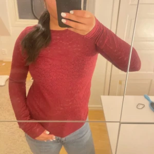 Vinröd blommig topp från KappAhl - Snygg vinröd långärmad topp från KappAhl i storlek S. Toppen har ett blommigt spetsmönster över hela och är tillverkad i ett mjukt, stretchigt material. Rund halsringning och normal passform, perfekt att styla med jeans eller kjol.