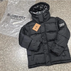 Svart pufferjacka från Burberry - Svart pufferjacka från Burberry med huva och klassiskt rutigt foder i grått, vitt och rött. Jackan har tryckknappar framtill, broderad logga på ärmen och är tillverkad i glansig polyester. Perfekt för kalla dagar och riktigt snygg streetstil.