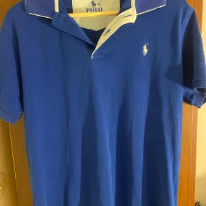 Blå pikétröja Polo Ralph Lauren XL - Klassisk blå pikétröja från Polo Ralph Lauren i 100% bomull. Tröjan har korta ärmar, vit broderad logga på bröstet och kontrastfärgad krage med vit insida. Perfekt för en stilren och sportig look.