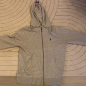 Grå hoodie från Polo Ralph Lauren - Grå hoodie från Polo Ralph Lauren med dragkedja framtill och klassisk huva med snörning. Tröjan har långa ärmar, två fickor och den ikoniska lilla Polo-loggan broderad på bröstet. Tillverkad i mjuk bomull för skön komfort.