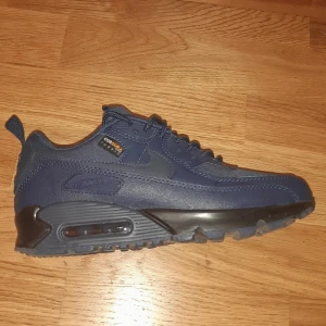 Nike Air Max 90 Surplus blå sneakers - Nike Air Max 90 Surplus sneakers i marinblått med svarta detaljer och en turkos accent framtill. Skorna har en robust platt sula med synlig Air Max-enhet, snörning och slitstark Cordura-överdel i syntet och textil. Perfekt för dig som gillar streetwear och sportig stil. Helt nya skor.