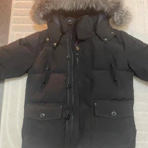 Svart dunjacka Moose Knuckles med päls - Säljer en svart dunjacka från Moose Knuckles med fet pälskant på huvan, stora fickor med knappar och dragkedja framtill. Jackan har klassisk logga på ärmen och broderad insida. Perfekt för kalla dagar och riktigt snygg streetstil.