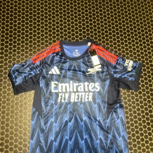 Arsenal bortatröja Adidas M blå - Arsenal bortatröja från Adidas i marinblått med blixtmönster i ljusblått och röda ränder på axlarna. Tröjan har klubbmärke och sponsortryck framtill, samt Aeroready-material som andas. Perfekt för dig som vill sticka ut på planen eller läktaren.