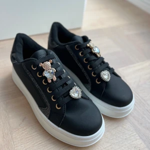 Svarta sneakers med strass och björn-detaljer - Svarta sneakers med vit platt sula, dekorerade med glittriga strassdetaljer i form av hjärtan och björnar på snörningen. Skorna har guldfärgade öljetter och svarta snören, samt glittriga svarta detaljer längs sidorna. Perfekta för dig som gillar att sticka ut.