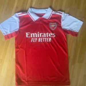 Arsenal röd och vit fotbollströja - Säljer en officiell Arsenal fotbollströja från Adidas med klassisk röd kropp och vita ärmar. Tröjan har vit krage med röda detaljer, klubbmärke på bröstet och Emirates Fly Better-tryck. Materialet är lätt och andas, perfekt för match eller träning.