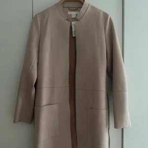Puderrosa kappa med bälte från H&M - Stilren puderrosa kappa från H&M i mjuk mocka-look med raka linjer och stora fickor framtill. Jackan har ett bälte i midjan och en enkel krage utan slag. Perfekt för dig som gillar en clean stil