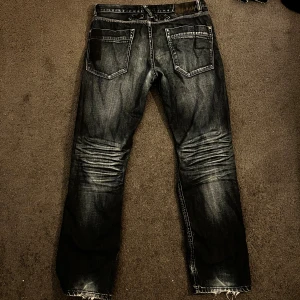 Svarta jeansbyxor med slitningar - Säljer ett par svarta jeansbyxor från kappahl med snygga slitningar och tvättade detaljer. Jeansen har raka ben, klassiska bakfickor och synliga sömmar. Materialet är robust denim och färgen är mörk med ljusare partier för en cool look. stort hål i skrevet som synns på bild 3-4 därav priset men kan du sy så har du fått ett fantastiska jeans! (passar ish (183-189