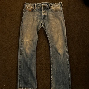 Diesel ljusblå jeans bootcut - Ljusblå Diesel jeans med bootcut passform, slitningar och hål på vänster ben. Klassiska femficksdetaljer, läderpatch bak och råa kanter vid benslut. Perfekta för dig som gillar en avslappnad och vintage look. Passar någon på 185 perfekt. litet hål vid knät som synns på bild