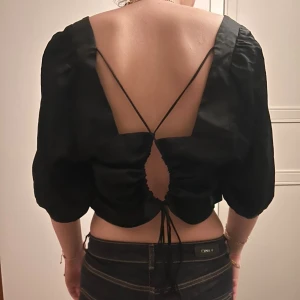 Svart croppad blus med öppen rygg - Även fast storleken är XL så passar den perfekt till nån som har XS-S vanligtvis. Säljer en svart croppad blus med fyrkantig urringning och trekvartsärmar. Baksidan har en stor öppen rygg med snörning och snygga knytband som ger en unik look. Materialet är glansigt och känns lyxigt, har ej kommit till användning 🥰🥰PRISET GÅR ATT BUDA PÅ🥰