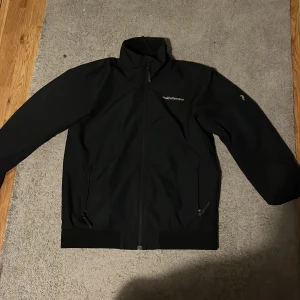 Svart vindjacka Peak Performance - Svart vindjacka från Peak Performance med stilren design och diskret logga på bröstet. Jackan har hög krage, varm,dragkedja framtill och två fickor med dragkedja. Perfekt för dig som gillar sportig och clean look.