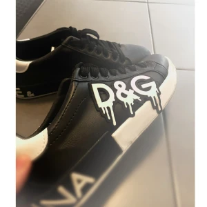 Svarta sneakers från Dolce & Gabbana - Säljer ett par svarta sneakers från Dolce & Gabbana med vit sula och coolt D&G-tryck i drip-style på sidan. Skorna har snörning, vita detaljer vid hälen och är tillverkade i skinn. Perfekta för dig som vill sticka ut med en unik streetstyle.