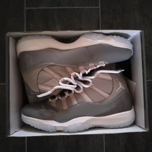 Nike Air Jordan 11 Cool Grey - Nike Air Jordan 11 Cool Grey med högt skaft, grå och vita detaljer samt vit sula. Skorna har snörning och en blandning av blankt och matt material, med ikoniska Jumpman-loggan på sidan. Perfekta för dig som vill sticka ut med en klassisk och stilren sneaker.