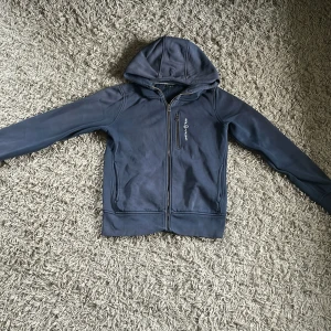 Blå hooded jacka från Sail Racing - Blå jacka med huva från Sail Racing, med dragkedja framtill och en extra bröstficka med zip. Jackan har lång ärm och är tillverkad i mjukt tygmaterial med fleecefoder på insidan. Snyggt broderad logga på bröstet och stilren design.