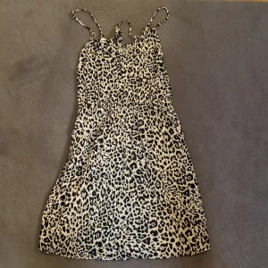 Leopardmönstrad kort klänning - Snygg kort klänning med leopardmönster och tunna axelband. Klänningen har en avslappnad passform och är perfekt för dig som vill sticka ut. Färgerna är svart och beige, vilket ger en cool kontrast. Materialet känns lätt och luftigt, perfekt för varma dagar.
