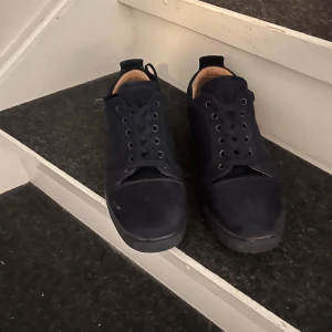 Svarta sneakers från Louboutin - Säljer ett par svarta sneakers från Christian Louboutin med klassisk röd sula. Skorna har snörning, rund tå och är tillverkade i mocka med skinnfoder. Perfekta för dig som vill ha en stilren och lyxig look med en ikonisk touch.