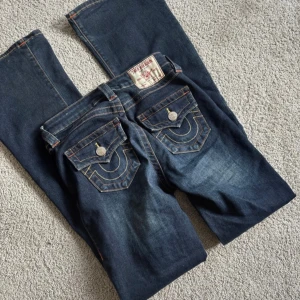 True Religion mörkblå bootcut jeans - Säljer ett par mörkblå jeans från True Religion med klassiska kontrastsömmar och ikoniska ficklock med silverknappar bak. Modellen har bootcut-ben och snyggt broderade detaljer på bakfickorna. Perfekt för dig som gillar statement-jeans med cool design.