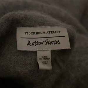 Grå polotröja från & Other Stories - Mjuk och fluffig grå polotröja från & Other Stories Stockholm Atelier i storlek S. Tröjan har lång ärm och en hög krage som ger en clean och chill vibe. Materialet är stickat och känns riktigt skönt mot huden. Perfekt för dig som gillar enkel och stilren look.
