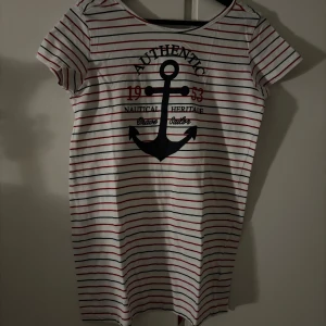 Randig kortklänning från Esmara - Säljer en vit kortklänning från Esmara med röda och marinblå ränder. Klänningen har ett stort ankare tryckt framtill och textdetaljer som ger en nautisk vibe. Rund halsringning och korta ärmar, tillverkad i mjuk bomull. Perfekt för dig som gillar marint tema.